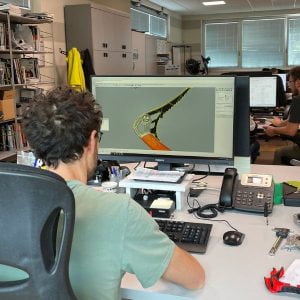 Ein Mann sitzt am Schreibtisch. Auf dem Bildschirm ist das CAD-Modell eines Eispickels zu sehen.