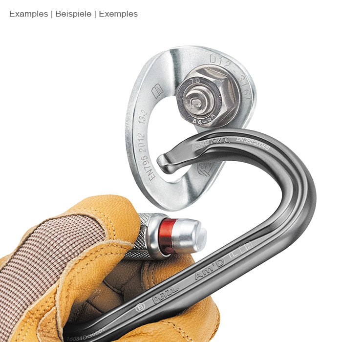 Coeur Stainless 12 von Petzl, silbernes Metallteil mit Karabinerhaken, gehalten von einer Hand mit gelbem Arbeitshandschuh und schwarzem Karabinerhaken mit rotem Verschlussmechanismus