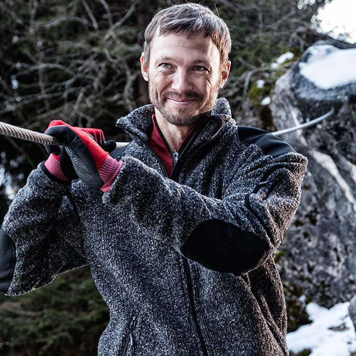 Mann trägt Grizzly Jacke graue Strickjacke mit schwarzem Besatz an Schultern und Ellbogen, rotem Unterziehshirt und roten Handschuhen, im Hintergrund Wald und Felsen mit Schnee
