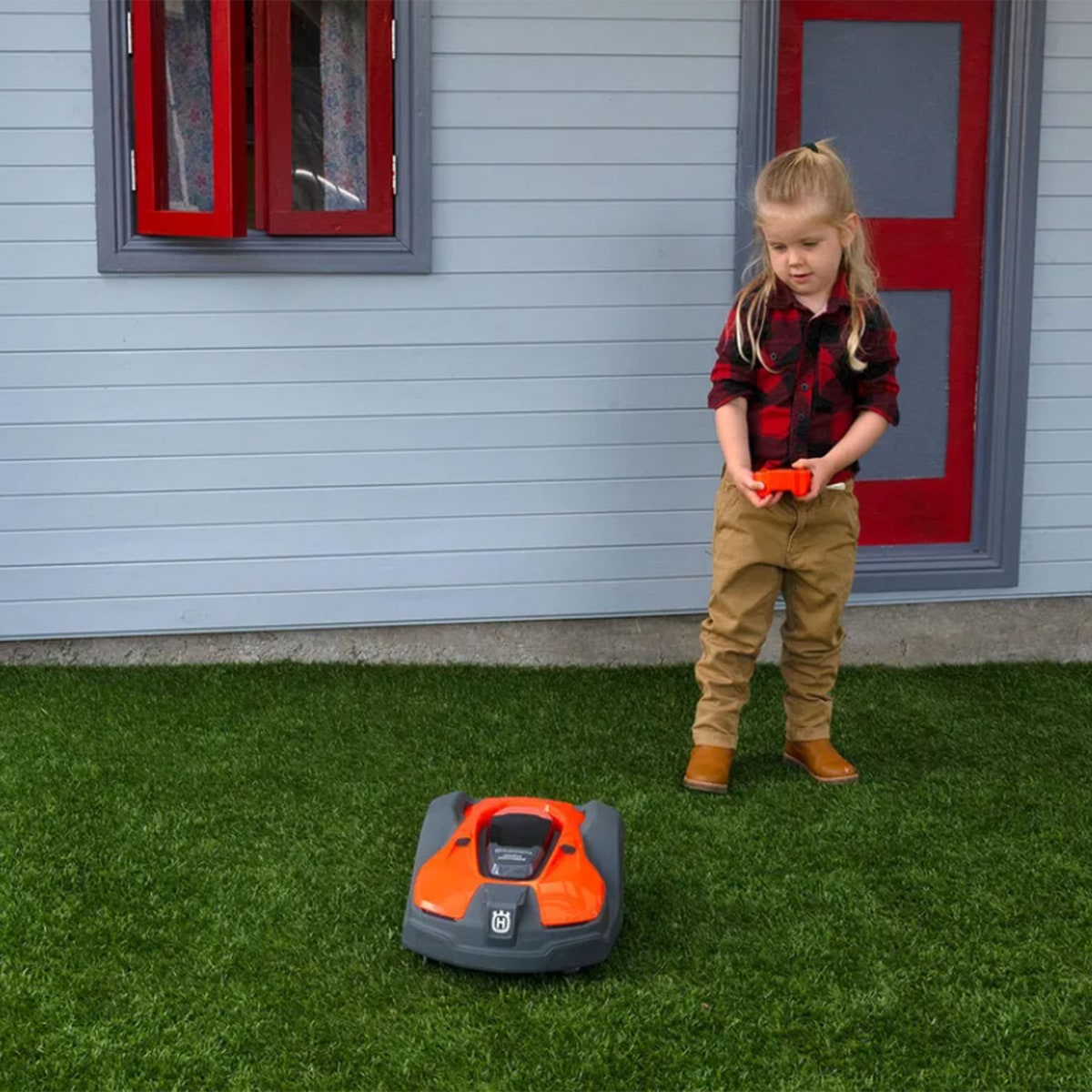 Ein Kind mit rotem kariertem Hemd, beigen Hosen und braunen Stiefeln steuert den orange-schwarzen Kinder Automower von Husqvarna auf grünem Rasen vor einem Haus mit rotem Fenster und Türrahmen