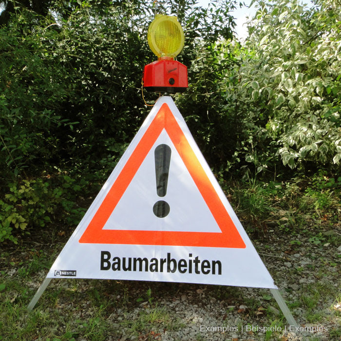 Warnfaltdreieck mit dem Aufdruck "Baumarbeiten" und einem Ausrufezeichen in einem orangefarbenen Dreieck, mit einer gelben Warnleuchte und rotem Gehäuse oben angebracht, auf Gras und Kies stehend