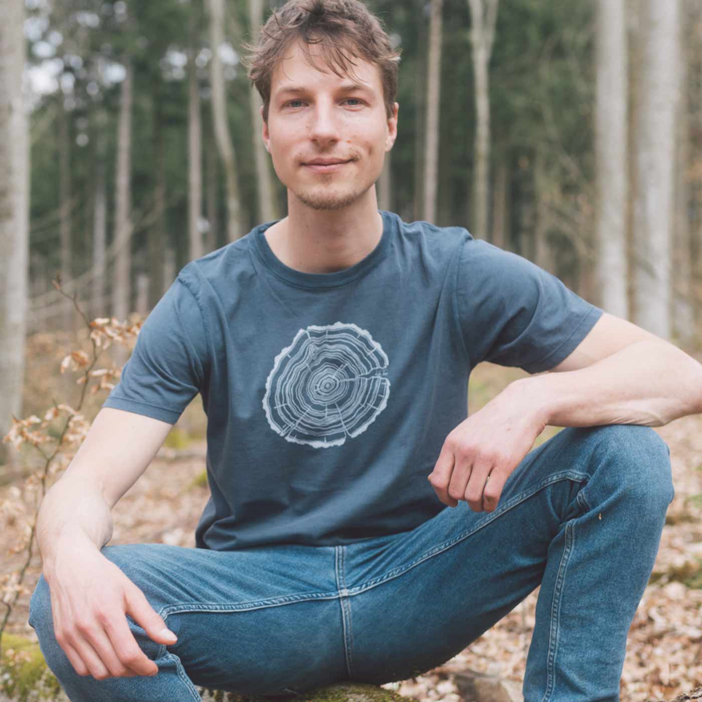 Ein Mann sitzt im Wald und trägt ein dunkelblaues T-Shirt mit einem weißen Baumstammschnitt, Produktname Treeslice