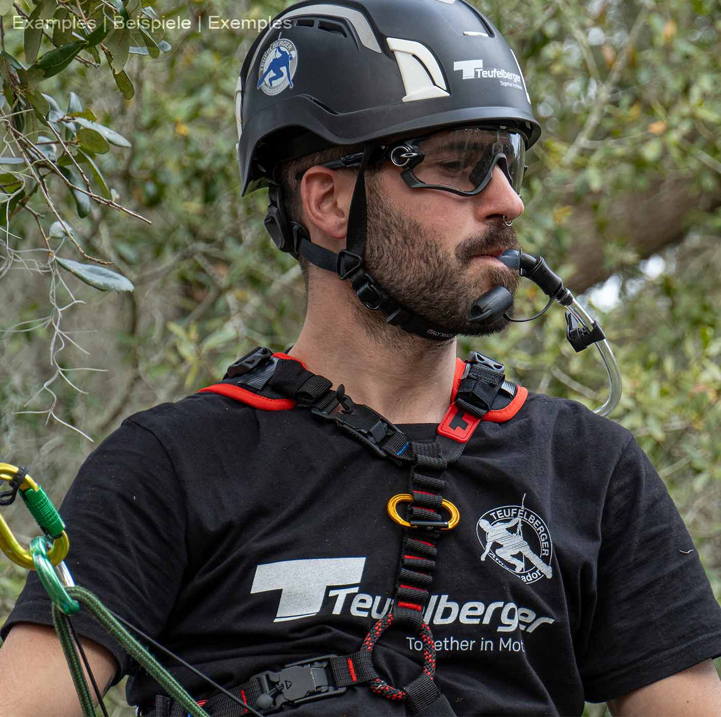 Mann trägt upMOTION SRT Klettergurt, schwarzen Helm und Schutzbrille, er hält einen Trinkschlauch im Mund, im Hintergrund Bäume und Blätter