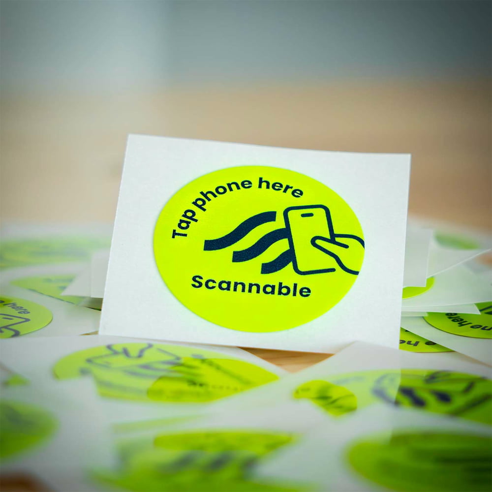 Nahaufnahme eines einzelnen runden, gelben Helm Sticker NFC Tag Aufklebers mit schwarzer Aufschrift „Tap phone here Scannable“ und einem Smartphone-Symbol, umgeben von weiteren Aufklebern