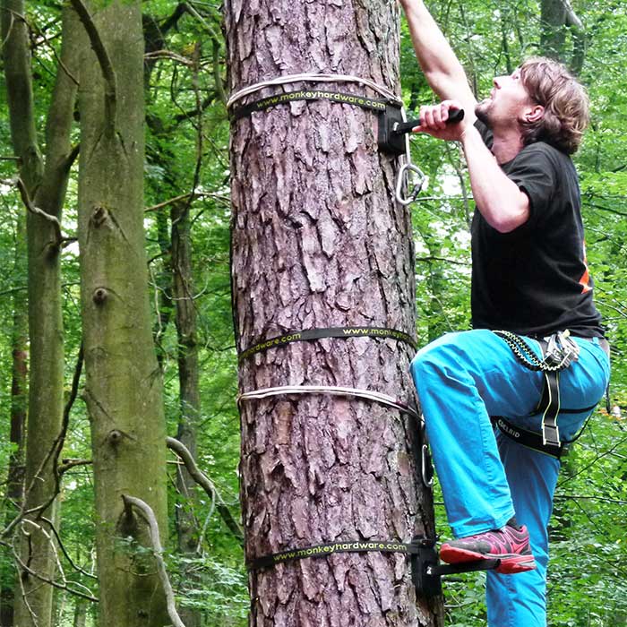 Tree-Monkey Steppers an Baumstamm befestigt, Mann mit blauem Hosenbein und schwarzem T-Shirt klettert mit Fuß auf Metallfuß und Gurt