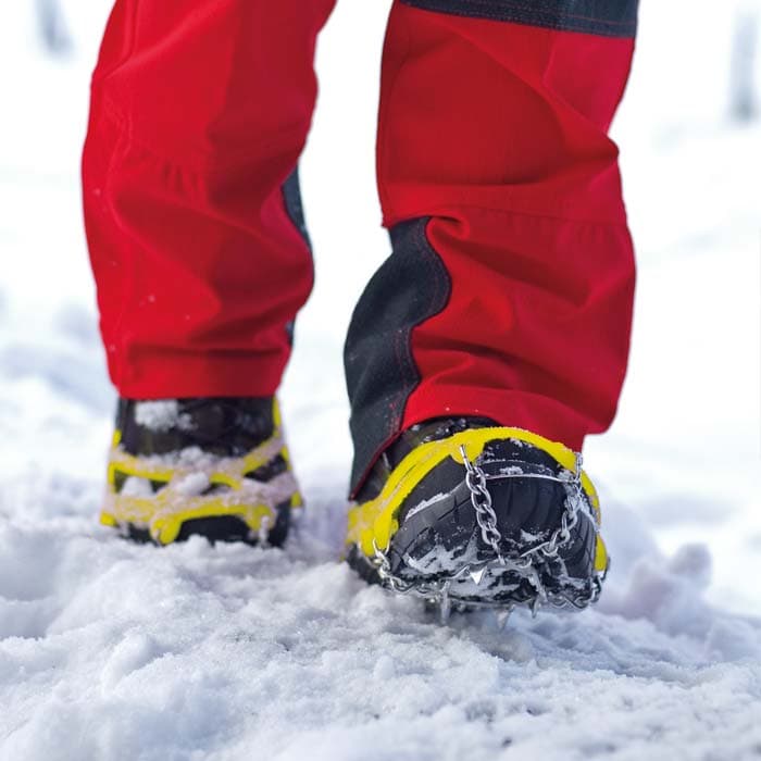 Spikes mit gelben Gummibändern und Metallketten an schwarzen Schuhen, getragen von einer Person mit roten Hosen im Schnee