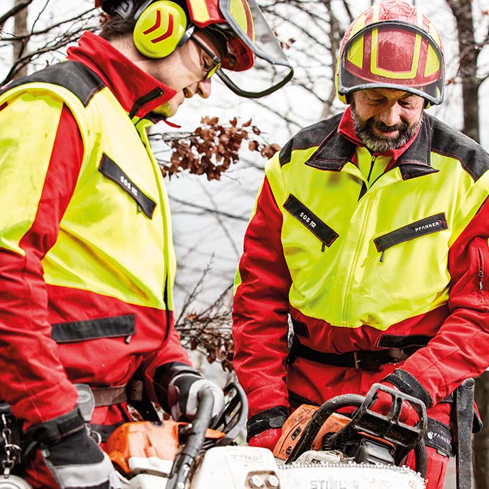 Zwei Personen tragen die KlimaAIR® Forstjacke in Gelb und Rot, eine Person hält eine Motorsäge, beide tragen Schutzhelme und Gehörschutz.
