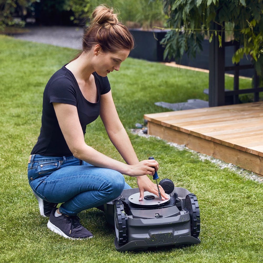 Frau kniet auf Rasen und repariert oder wartet 310 Mark II Mähroboter mit Schraubendreher, Gartenumgebung sichtbar