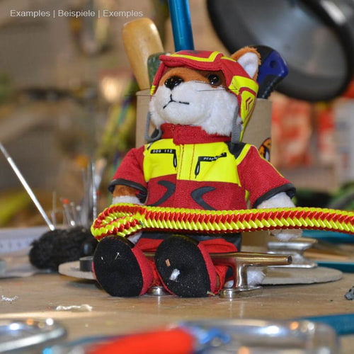 Wiffzack Plüschfigur sitzt auf einem Tisch, umgeben von Werkzeugen, trägt rote und gelbe Kleidung und Helm