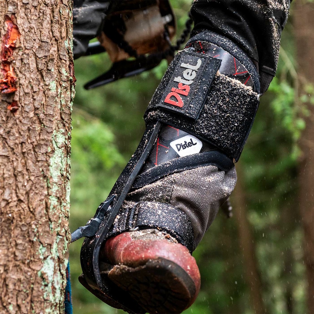 Distel Carbon 3.1 Velcro von Distel, Klettersteigsporn am Bein eines Trägers, der einen Baumstamm berührt, mit sichtbarem Schmutz auf dem Sporn und Schuh