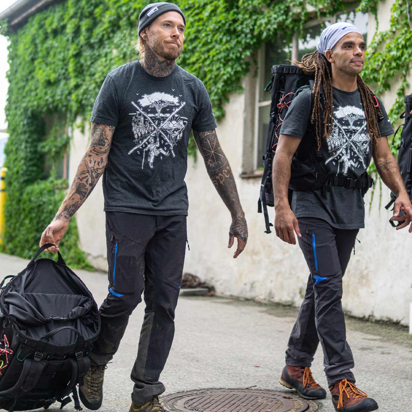 Zwei Männer tragen das dunkelgraue tSPIRIT T-Shirt mit weißem Kletter- und Naturmotiv, einer trägt eine schwarze Mütze und hält eine Tasche, der andere trägt ein helles Kopftuch und einen Rucksack