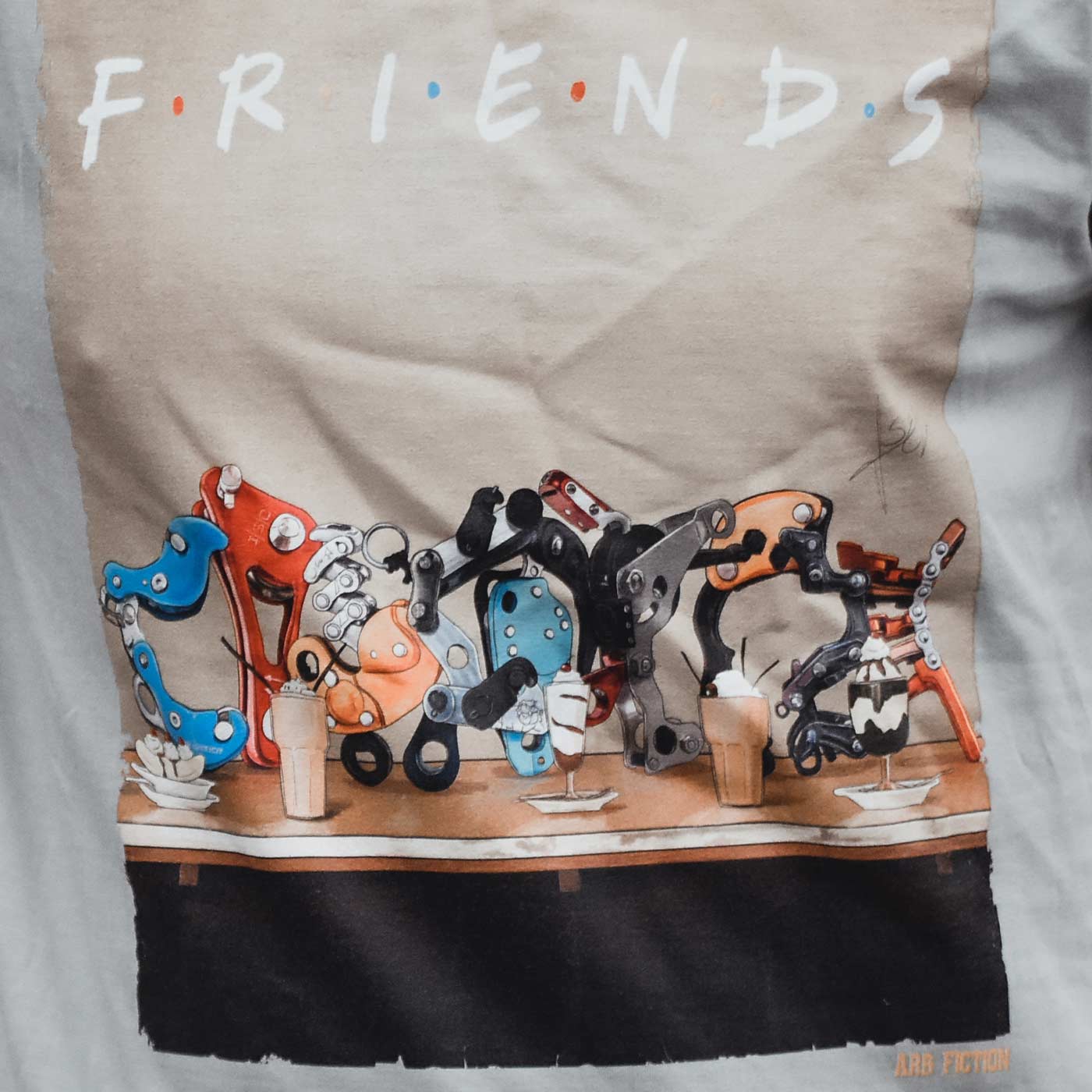 Nahaufnahme des T-Shirt-Aufdrucks mit dem Schriftzug "Friends" und detaillierten mechanischen Teilen in verschiedenen Farben auf einem Tisch, unten rechts der Schriftzug "ARB FICTION" sichtbar