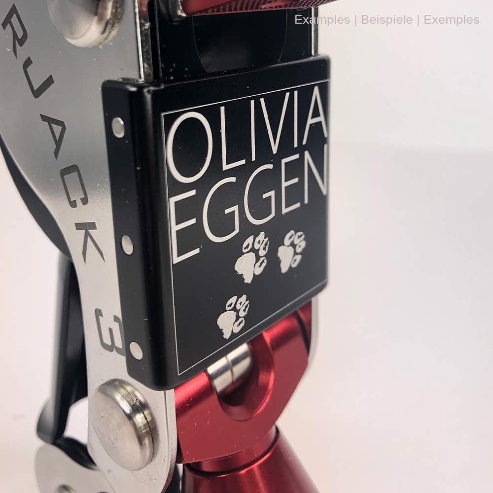 Nahaufnahme des schwarzen Schilds am SpiderJack 3 mit weißer Gravur "OLIVIA EGGEN" und Pfotenabdrücken, roter Metallhaken und silberner Rahmen