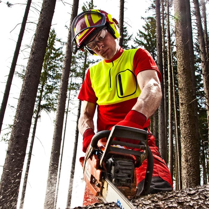 Mann trägt das Zipp-Neck Shirt in rot und gelb, Schutzhelm, Schutzbrille und Handschuhe, hält eine Motorsäge im Wald