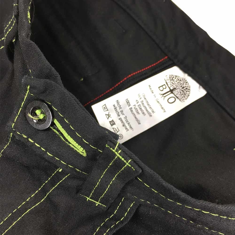 Nahaufnahme des Bunds der Roots Pure Outdoorhose in Schwarz mit grünem Knopfloch und grünem Faden, innen ein weißes Etikett mit Text und Baumlogo