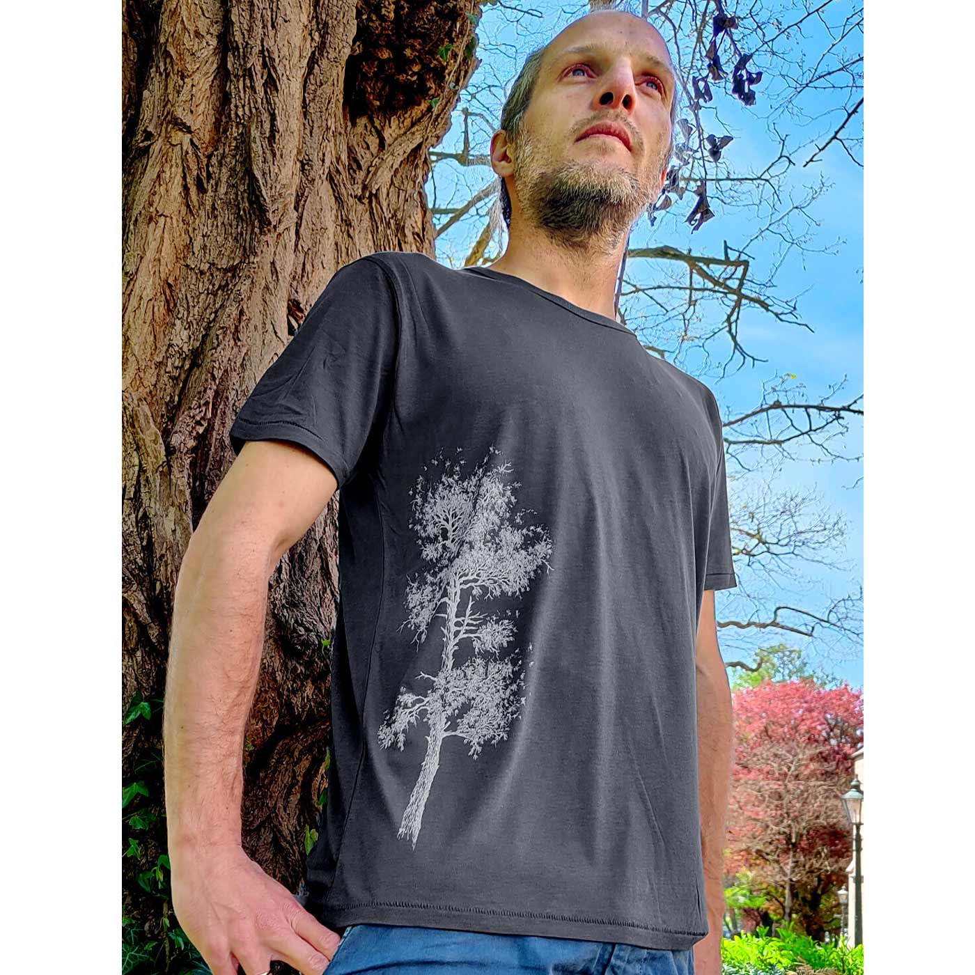 Mann steht draußen vor einem Baum und trägt ein dunkelgraues Pine Tree T-Shirt mit einem weißen Baumdruck auf der linken Seite des Shirts.