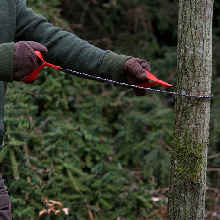 Person mit grüner Jacke und Handschuhen hält die Nordic Pocket Saw mit roten Schlaufen um einen Baumstamm gespannt