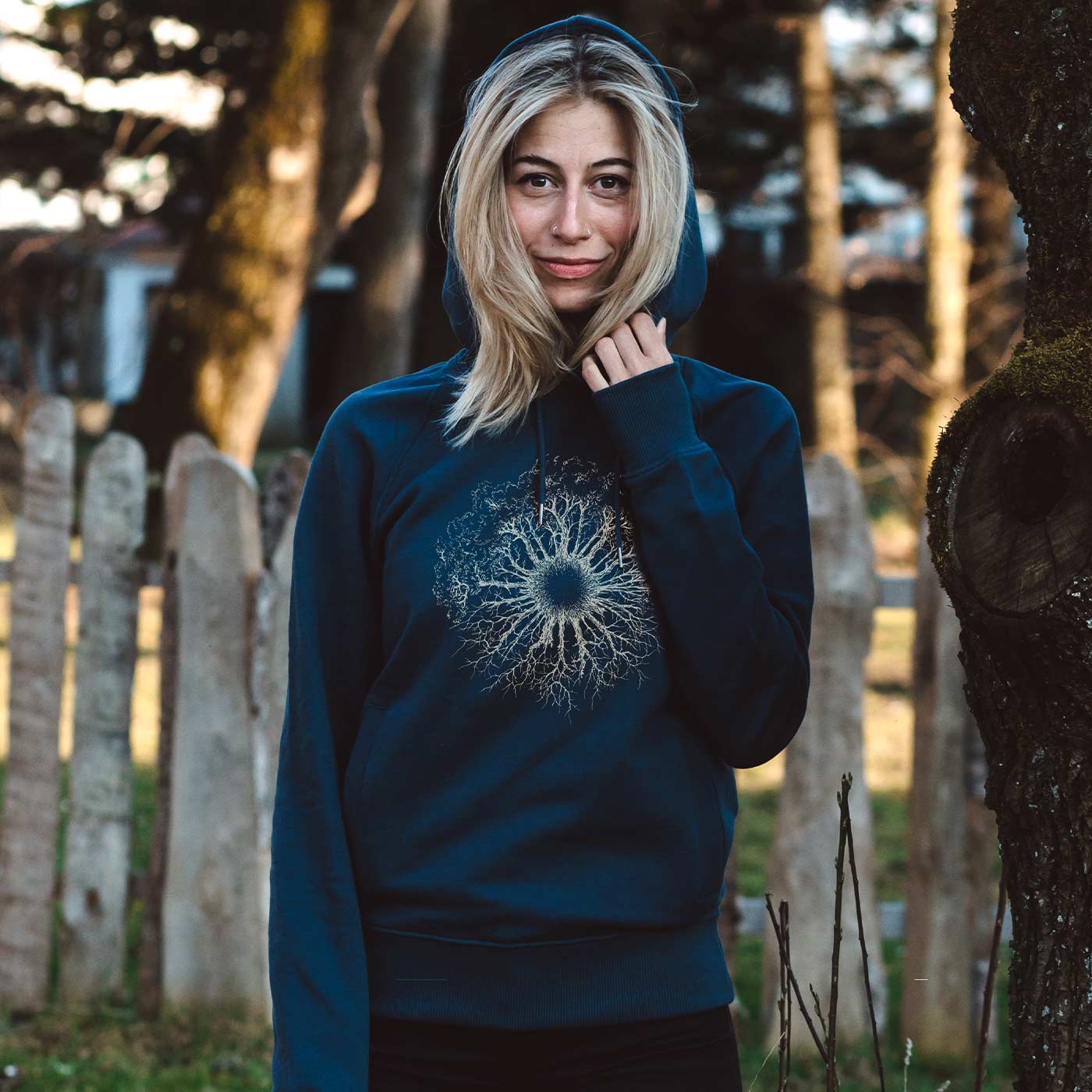 Eine Frau steht draußen neben einem Baum und trägt einen blauen Wooden Iris Hoodie mit Kapuze und einem weißen, baumwurzelartigen Muster auf der Brust.