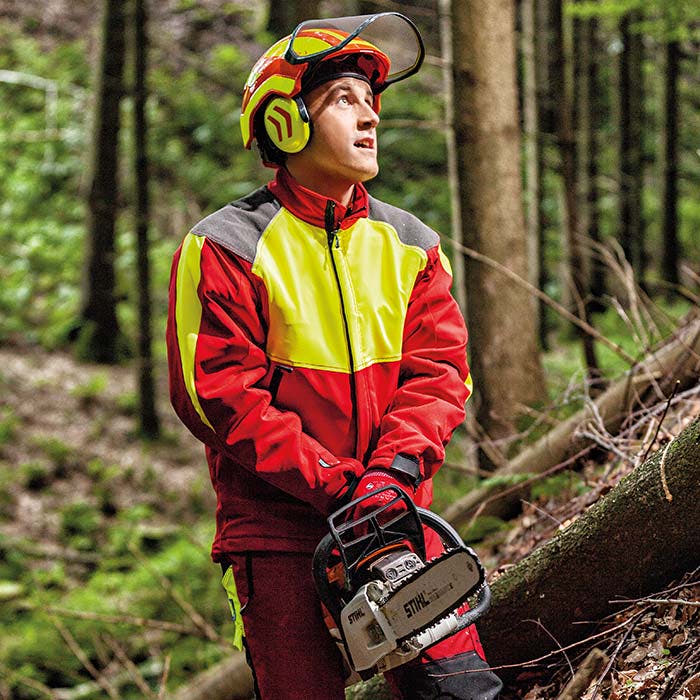 Mann trägt Schnittschutzjacke in Rot mit gelben und grauen Akzenten, dazu Helm und Handschuhe, hält eine Motorsäge im Wald