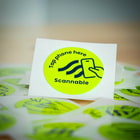 Nahaufnahme eines einzelnen runden, gelben Helm Sticker NFC Tag Aufklebers mit schwarzer Aufschrift „Tap phone here Scannable“ und einem Smartphone-Symbol, umgeben von weiteren Aufklebern