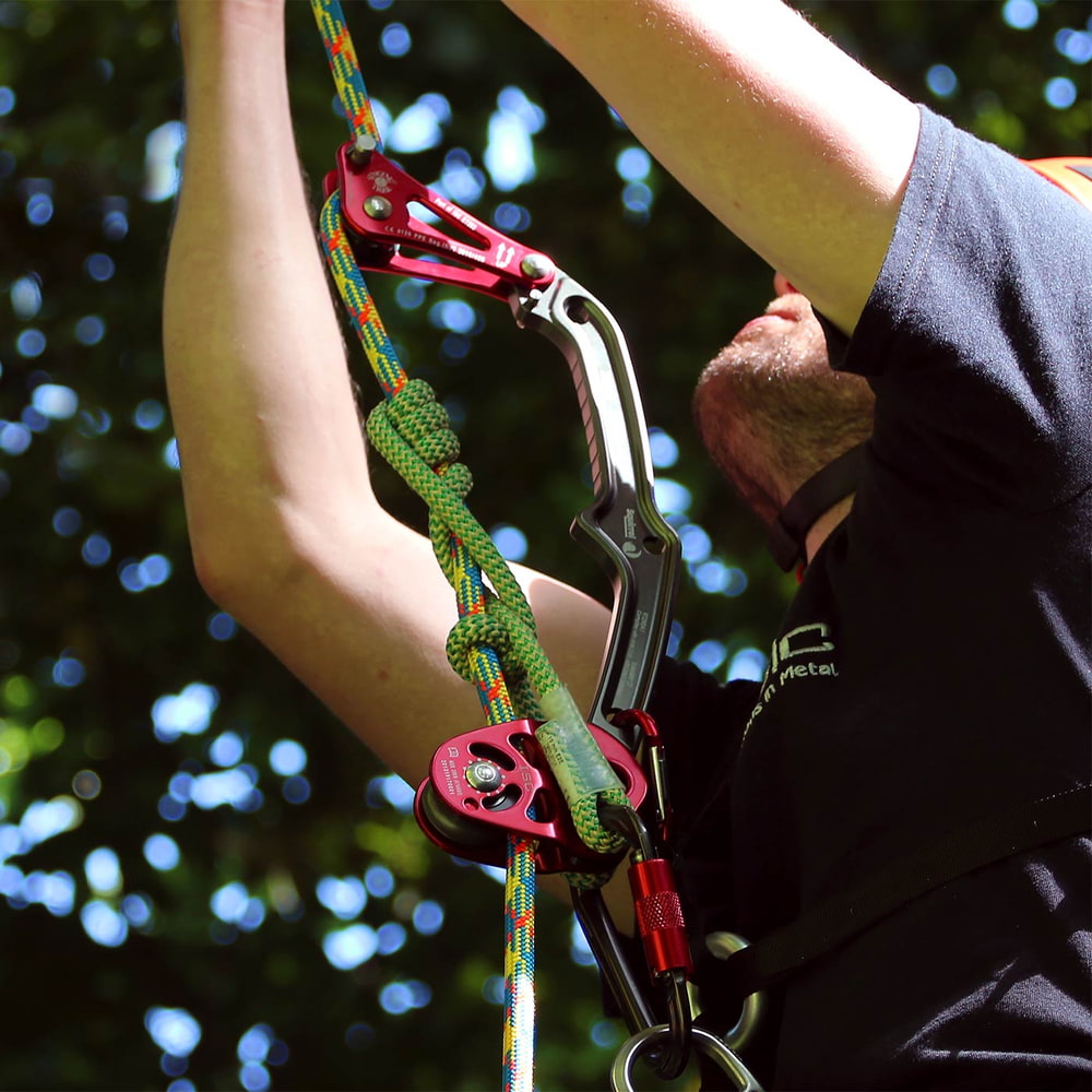 Person mit schwarzem Shirt hält Rope Wrench Squirrel PPE Regulations System Kit mit rotem und schwarzem Metallgerät, grün-weißem Seil und schwarzem Karabiner von ISIC in der Hand, Hintergrund grüne Blätter und Himmel