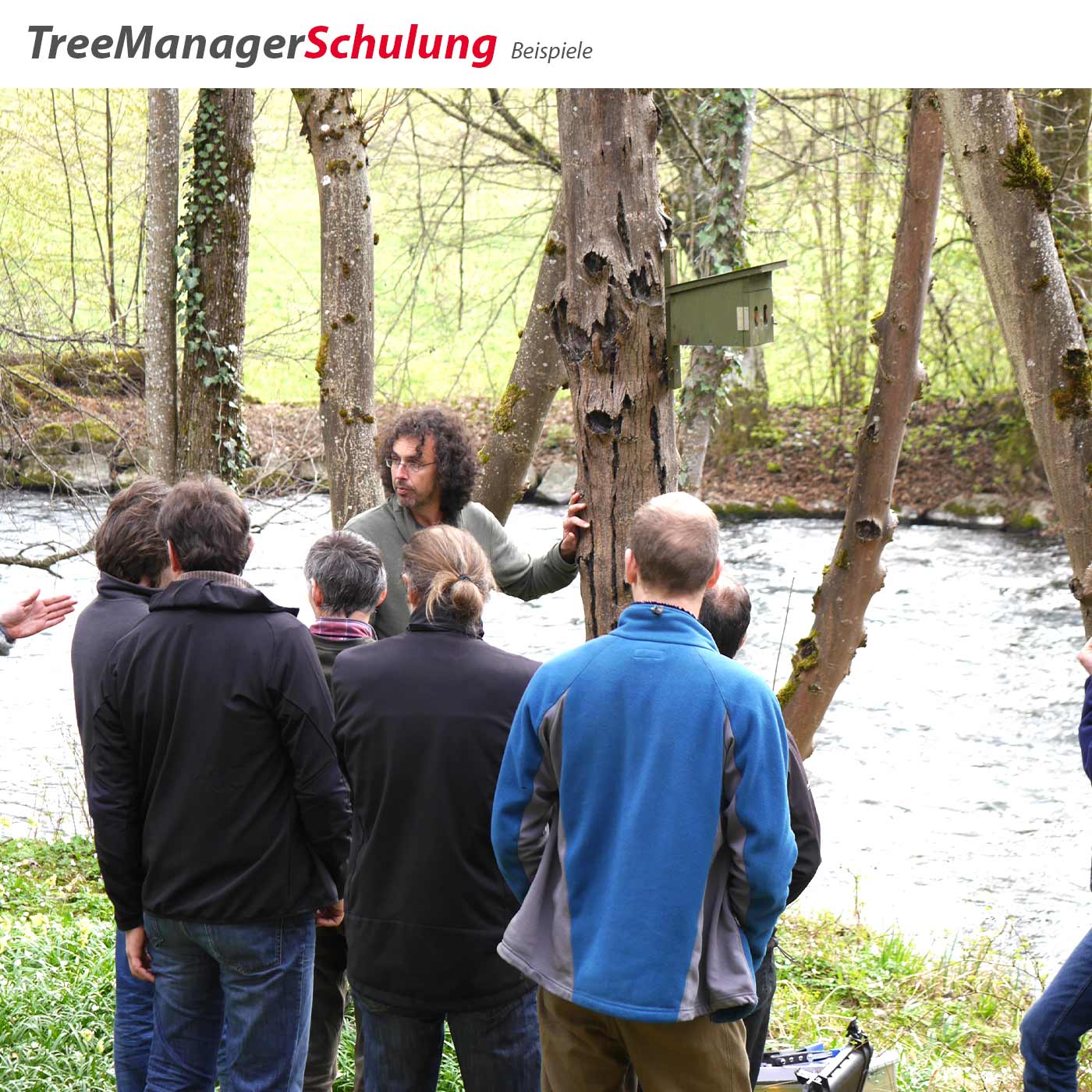 Gruppe von sieben Personen, einer zeigt an einem Baum mit einem Kästchen, im Hintergrund ein Fluss und Bäume