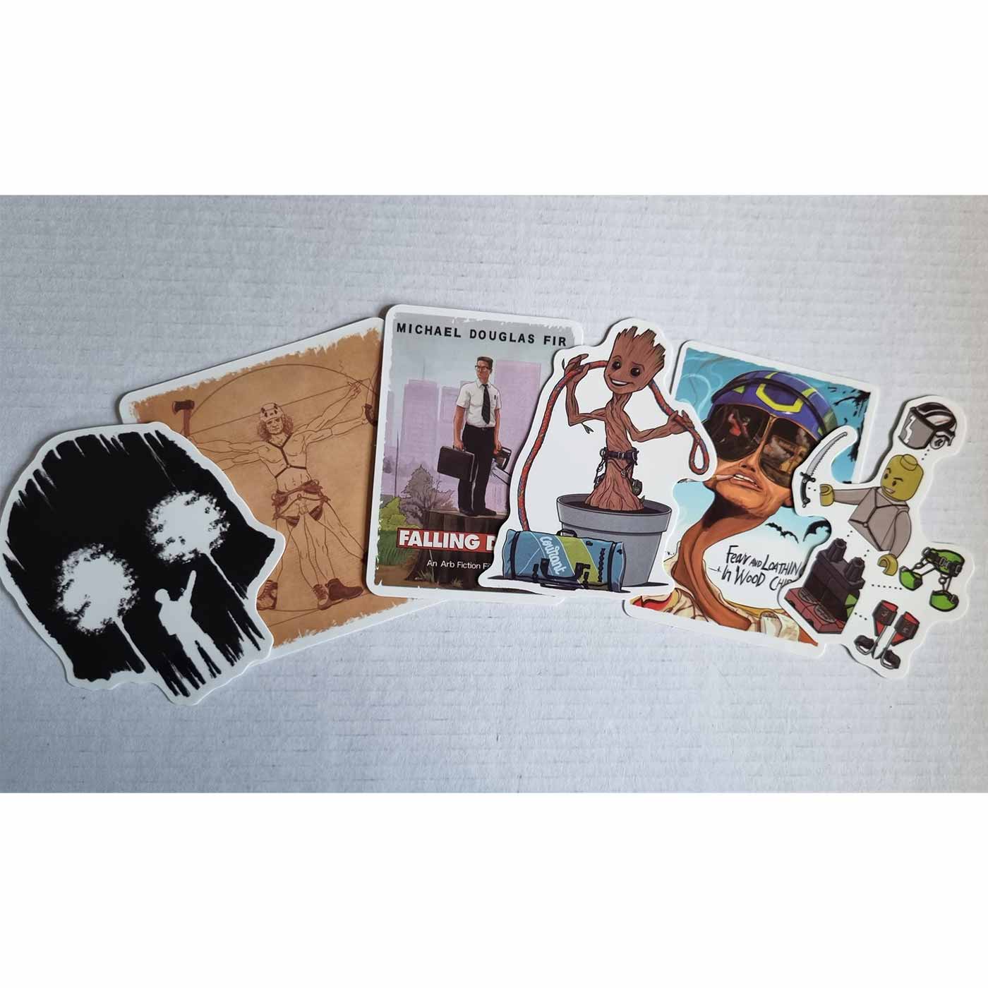 Sechs ARB Fiction Pack Vol. 3 Aufkleber liegen nebeneinander auf weißem Hintergrund, darunter ein schwarzer Aufkleber mit zwei weißen Bäumen, ein Aufkleber mit einem Mann in Anzug und Kettensäge mit der Aufschrift "Michael Douglas Fir Falling Down", ein t