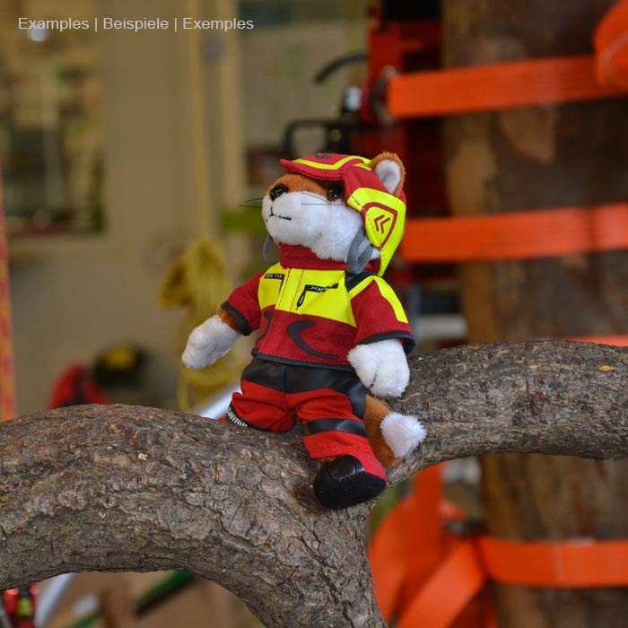Wiffzack Plüschfigur sitzt auf einem Baumstamm, trägt rote und gelbe Kleidung und Helm, im Hintergrund Werkstattumgebung sichtbar