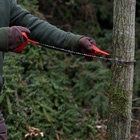 Person mit grüner Jacke und Handschuhen hält die Nordic Pocket Saw mit roten Schlaufen um einen Baumstamm gespannt