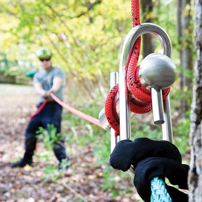 Port A Wrap Large aus glänzendem Metall, an dem rote und blaue Seile befestigt sind, im Wald, mit einer Person im Hintergrund, die ein Seil hält.