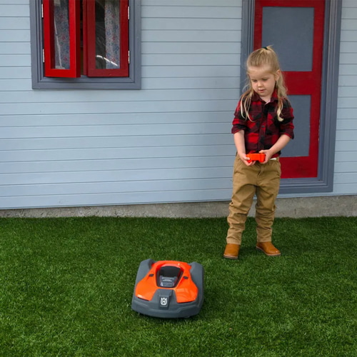 Ein Kind mit rotem kariertem Hemd, beigen Hosen und braunen Stiefeln steuert den orange-schwarzen Kinder Automower von Husqvarna auf grünem Rasen vor einem Haus mit rotem Fenster und Türrahmen