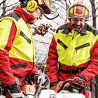 Zwei Personen tragen die KlimaAIR® Forstjacke in Gelb und Rot, eine Person hält eine Motorsäge, beide tragen Schutzhelme und Gehörschutz.