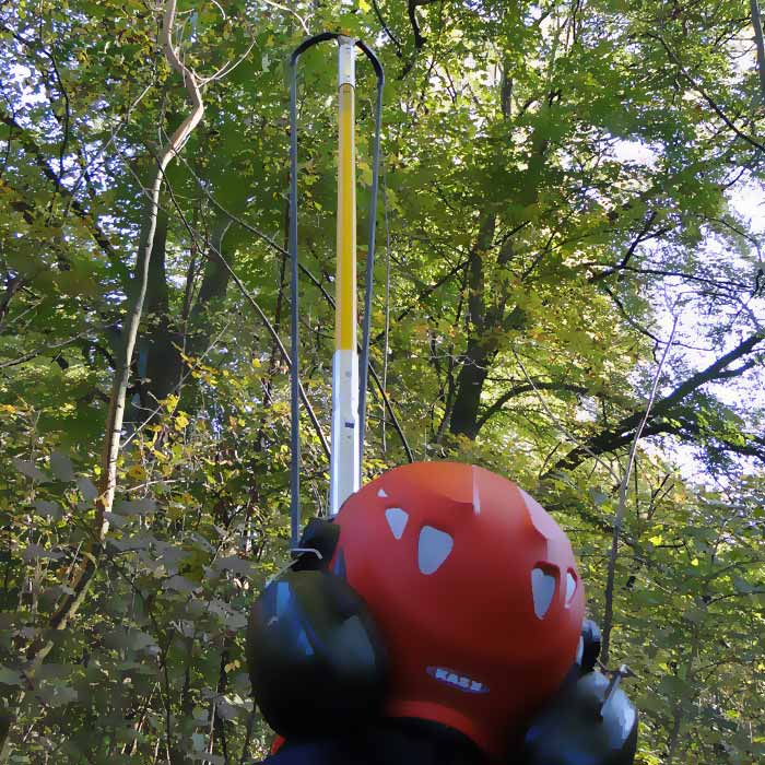 Person mit rotem Helm und Gehörschutz von hinten, hält den Big Shot Stab mit schwarzem Griff und gelbem Gurtband in einem Wald