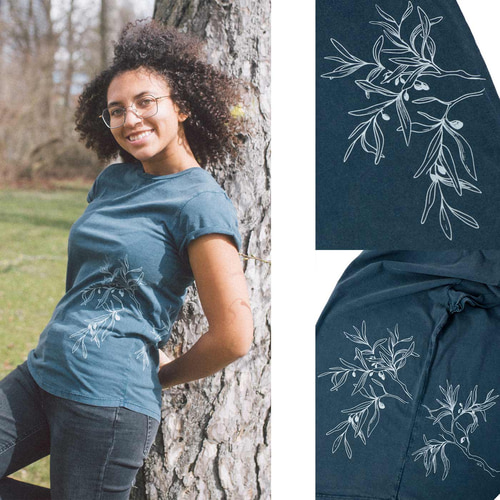 Eine Frau lehnt an einem Baum und trägt ein dunkelblaues T-Shirt "Olive Branch" mit weißem Olivenzweig-Motiv, daneben zwei Nahaufnahmen des Drucks.