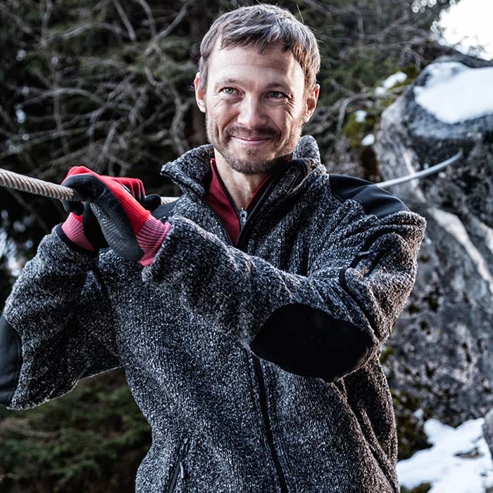 Mann trägt Grizzly Jacke graue Strickjacke mit schwarzem Besatz an Schultern und Ellbogen, rotem Unterziehshirt und roten Handschuhen, im Hintergrund Wald und Felsen mit Schnee