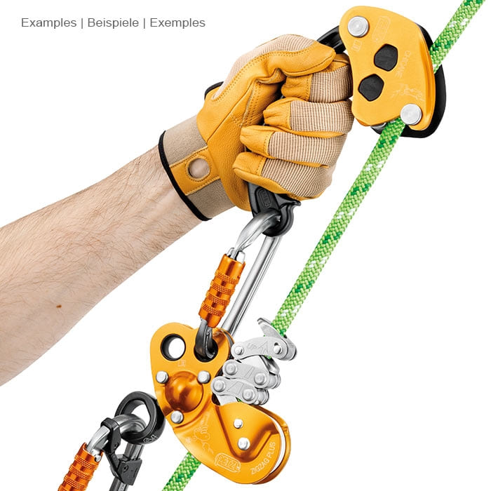 Petzl Chicane, gelb-schwarzes Sicherungsgerät mit Griff, gehalten von einer Hand mit gelbem Handschuh, verbunden mit einem Karabiner und einem gelben Sicherungsgerät an einem grünen Seil